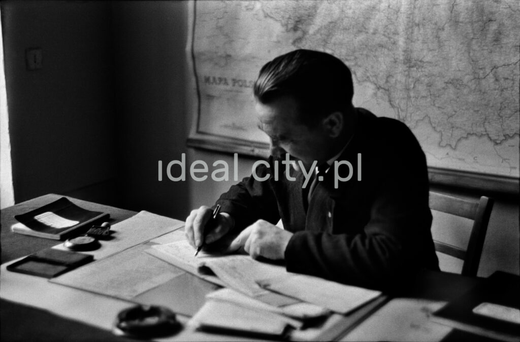 A man in a jacket is writing something bent over a desk, behind him a map hangs on the wall.