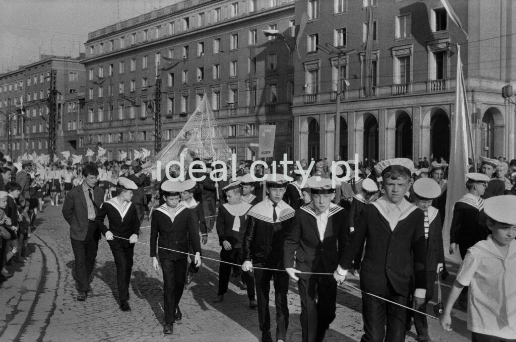 Boys in sailor's clothes march along a wide avenue with apartment blocks in the background.