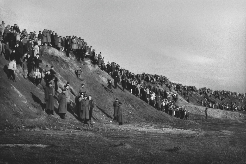 The crowd of people on the slope of a long escarpment looks in one direction.