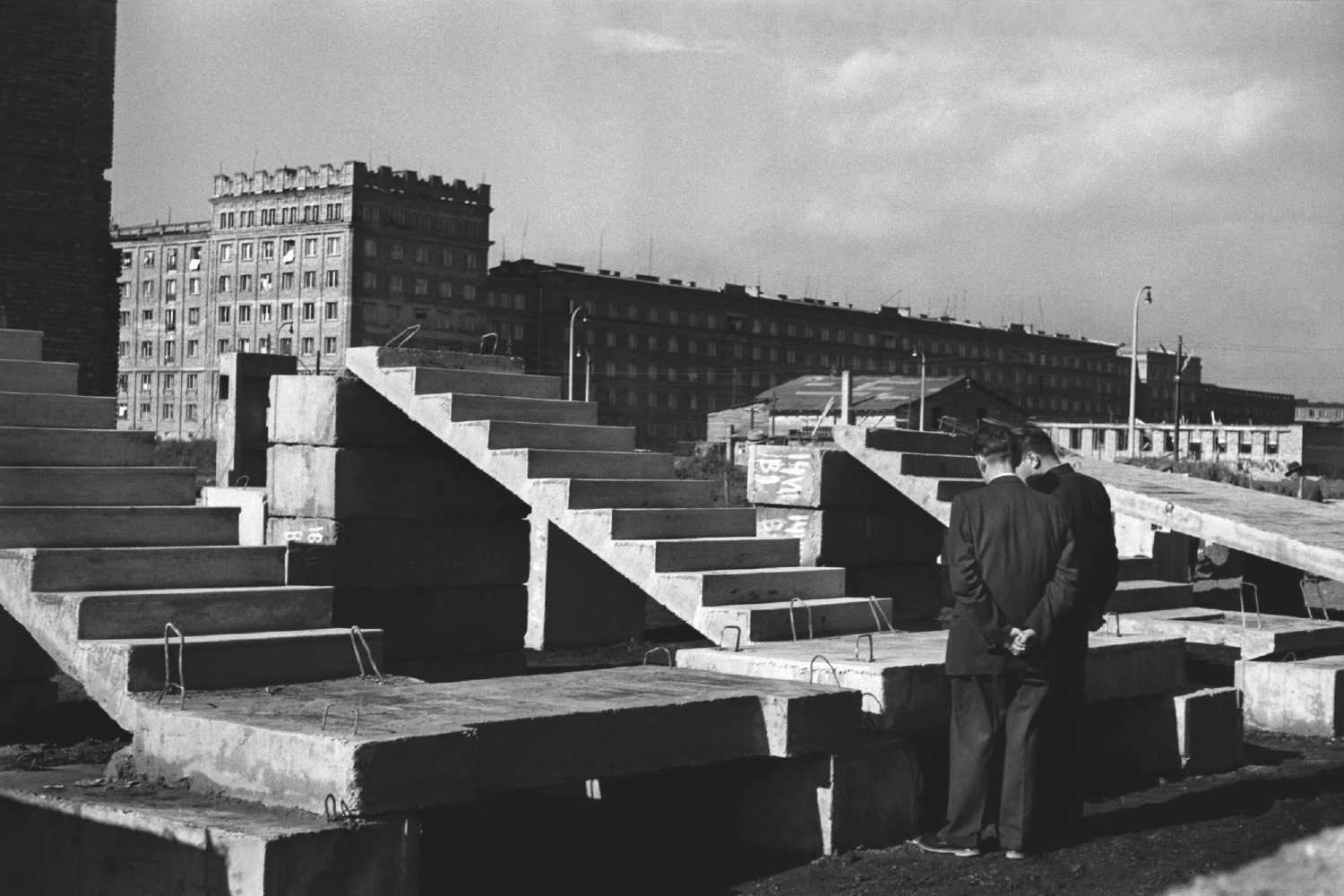 Two men in suits look at types of prefabricated stairs in public view. Residential buildings in the background.