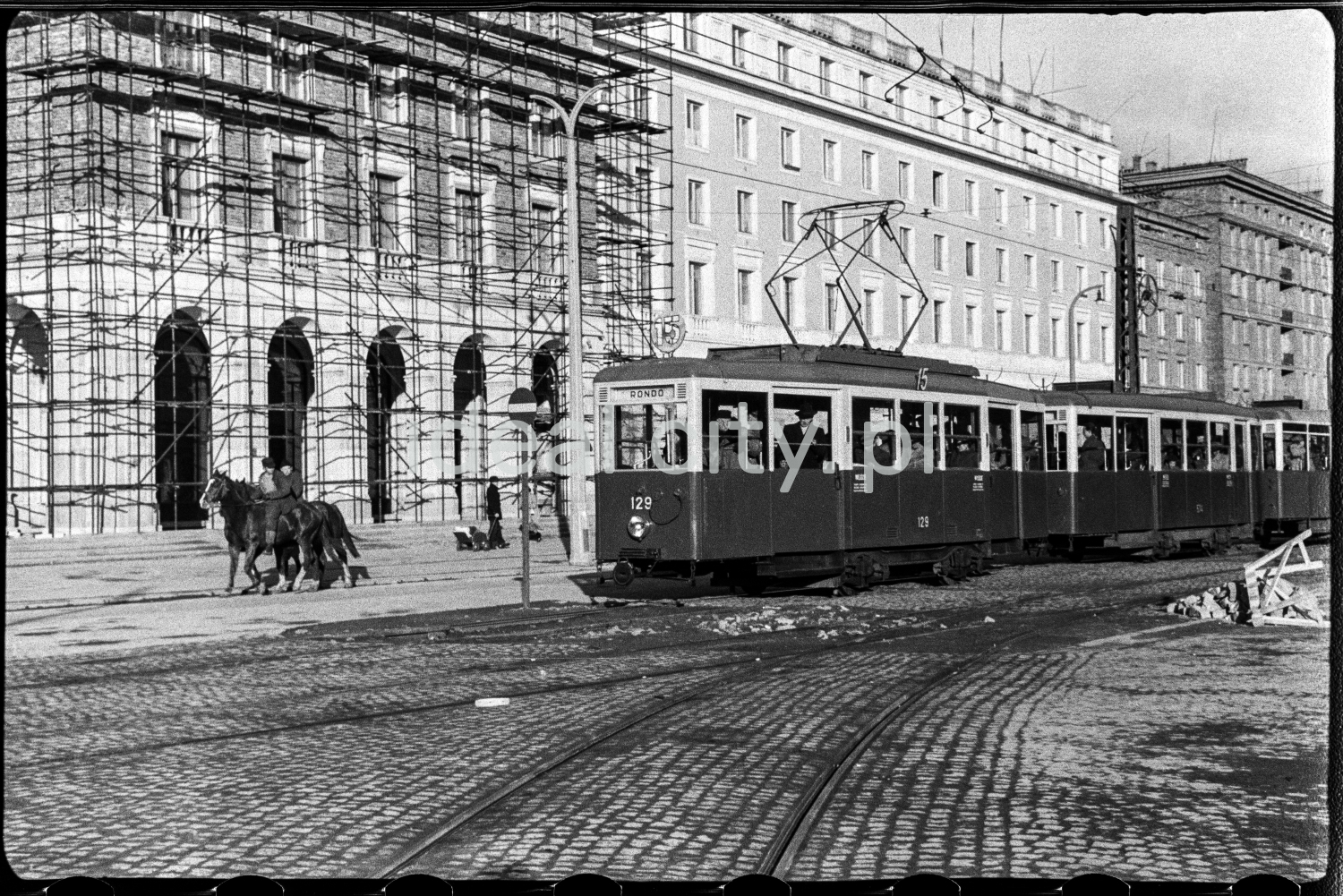Wybrukowaną aleją jedzie tramwaj oraz jeździec na koniu, w tle finałowe prace tynkarskie przy socrealistycznej zabudowie mieszkalnej.
