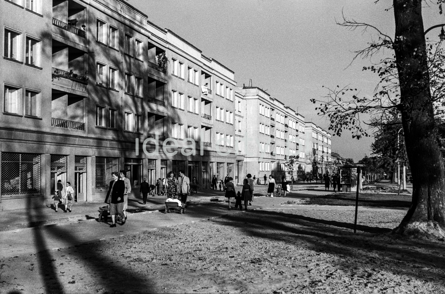 Ruch pieszy na szerokim chodniku przed ciągiem trzypiętrowych bloków mieszkalnych.