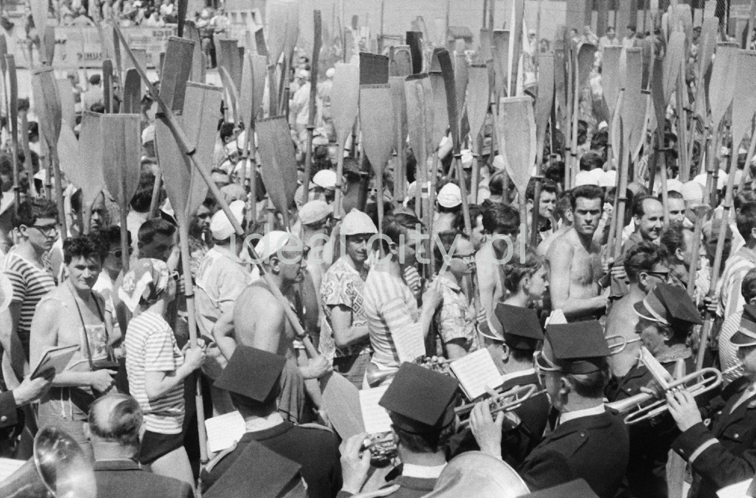 Tłum ludzi stoi z podniesionymi na sztorc wiosłami.