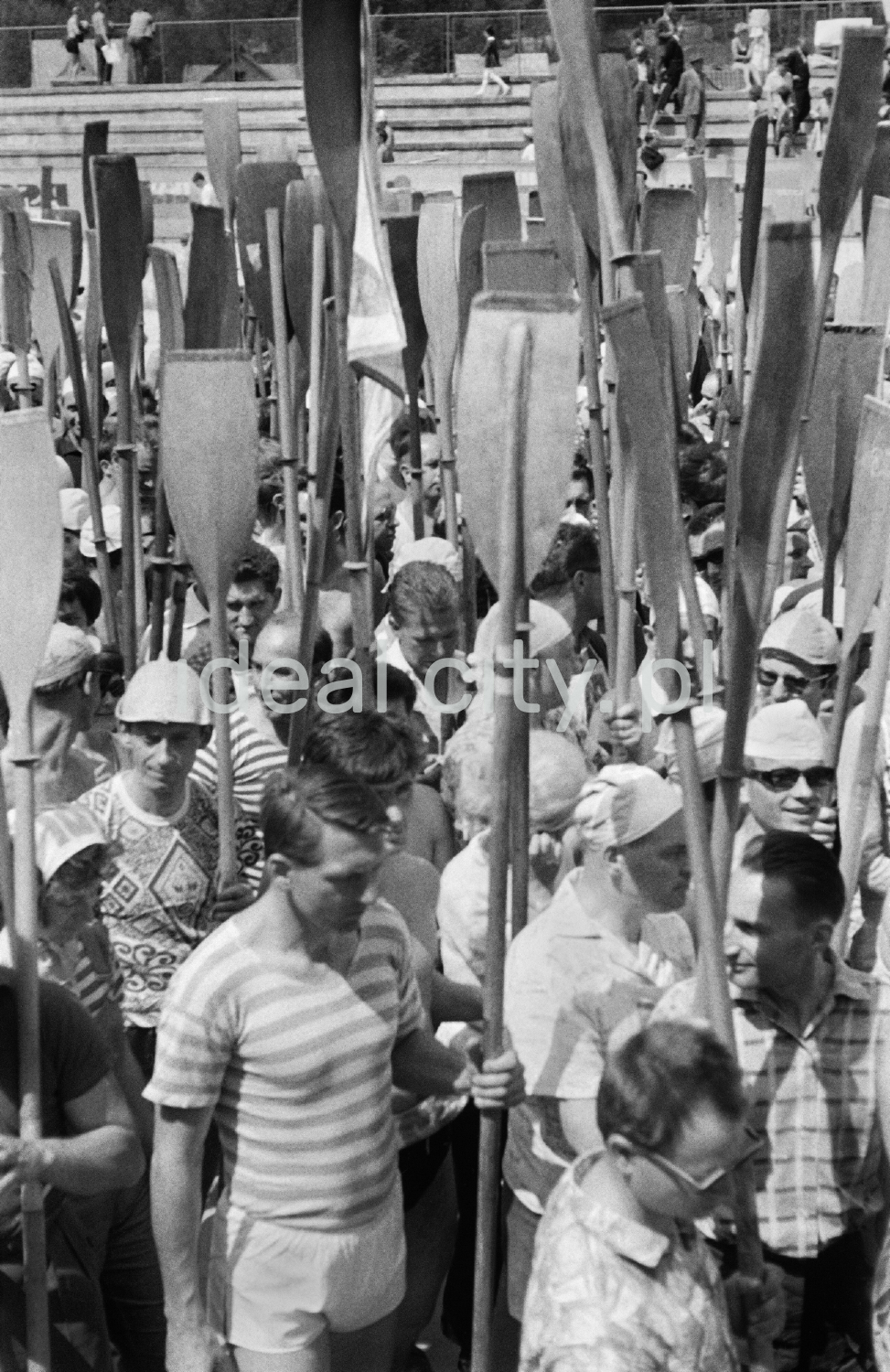 Tłum ludzi stoi z podniesionymi na sztorc wiosłami.