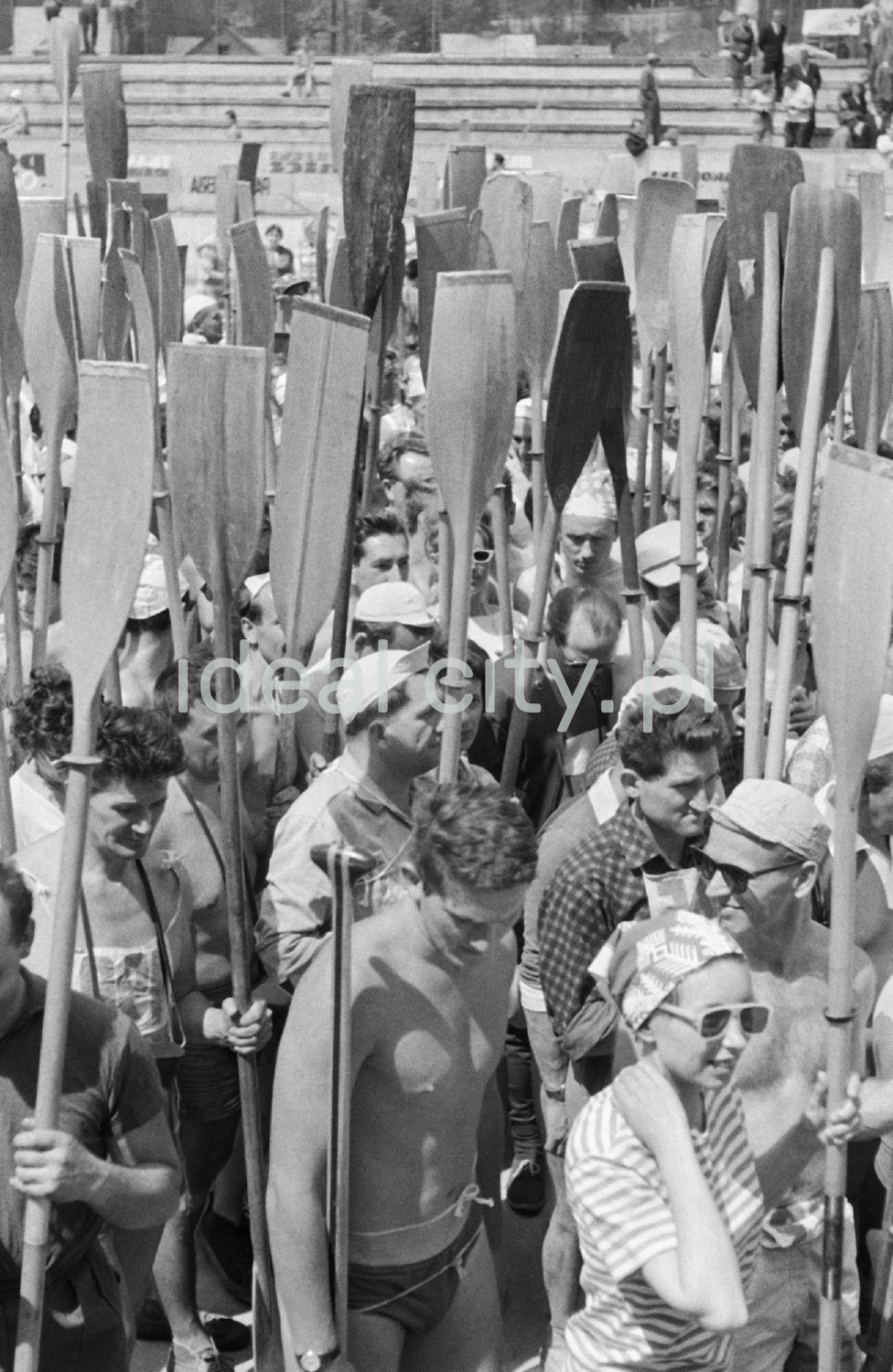 Tłum ludzi stoi z podniesionymi na sztorc wiosłami.