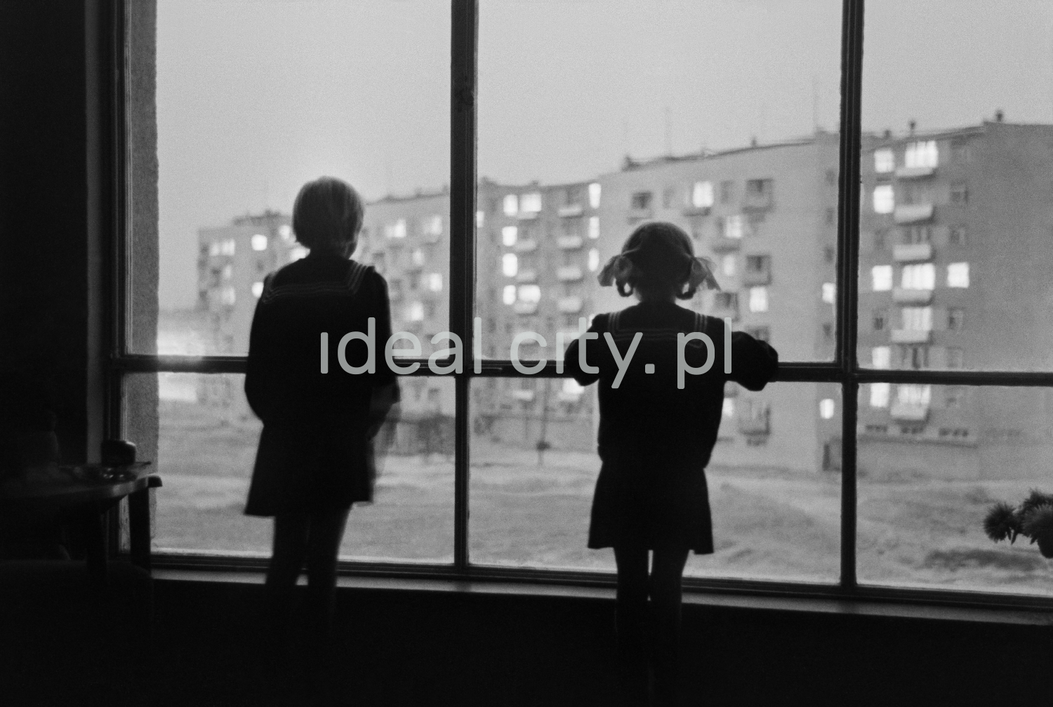 Kontury dwójki dzieci spoglądających z dużego okna na piętrze bloku mieszkalnego w stronę podwórka osiedla.