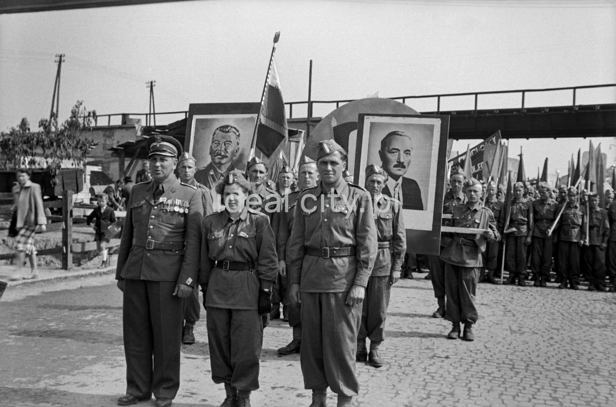 Mężczyźni i kobiety w mundurach maszerują w pochodzie, nad tłumem portrety Stalina i Bieruta.