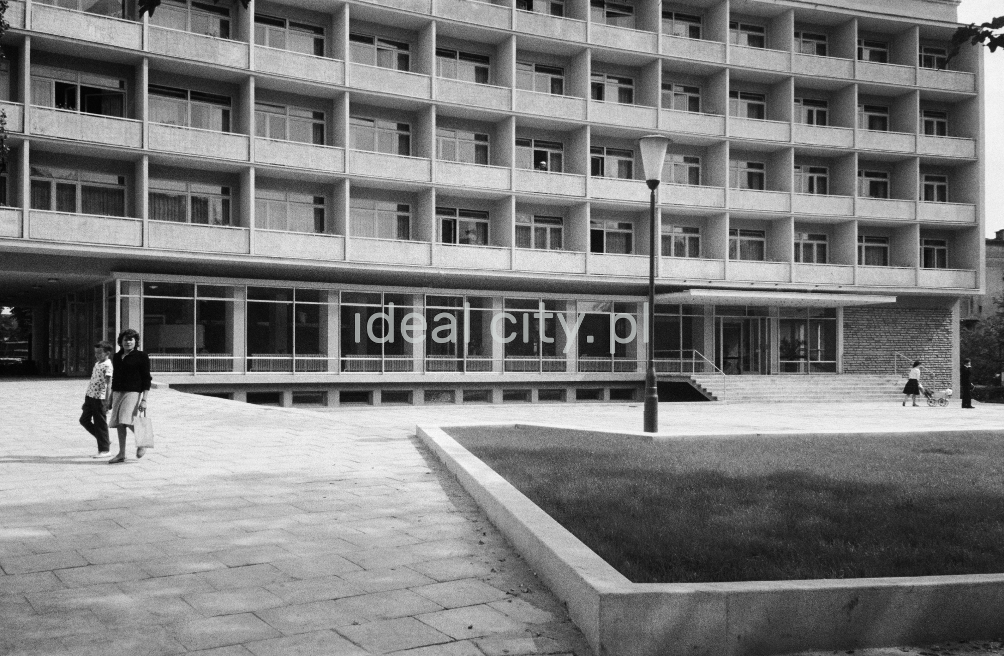 Matka z dzieckiem przechodzi przed bryłą modernistycznego hotelu.