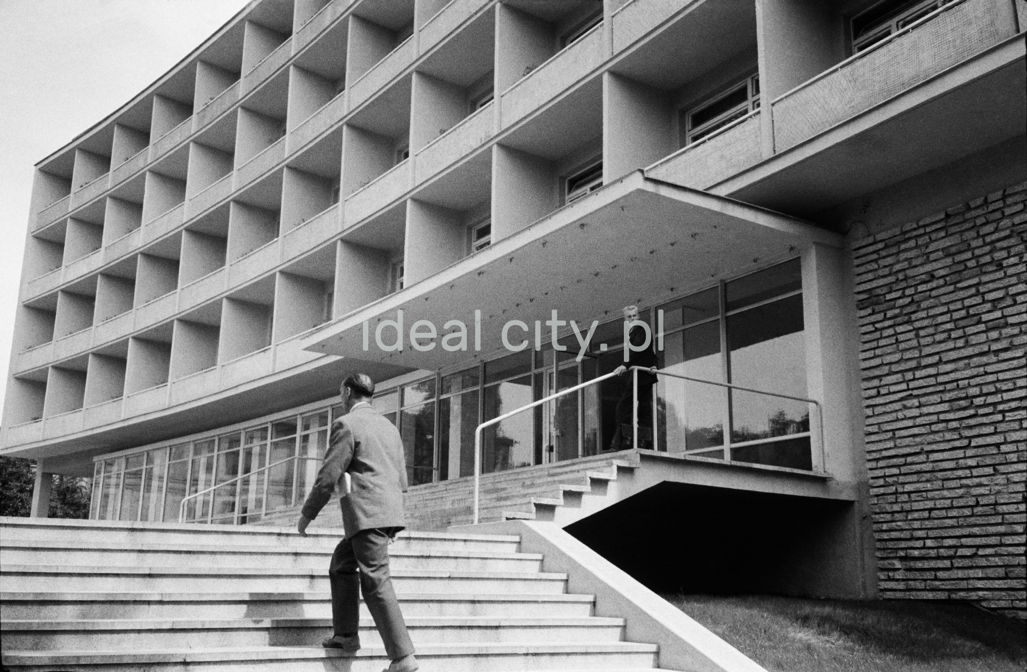 Mężczyzna w garniturze wychodzi po schodach przed modernistyczną bryłą hotelu.