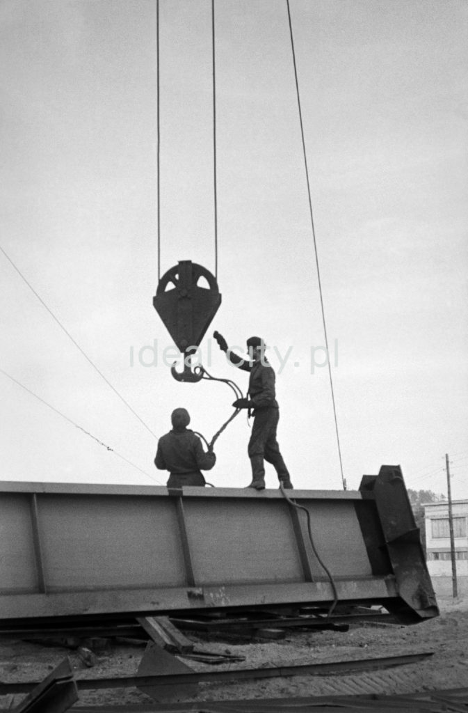 Dwóch robotników montuje linę na zwisającym z góry potężnym haku podnośnika.