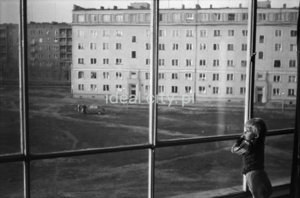 Dziecko spogląda na podwórko pomiędzy blokami przez obszerne okno swojego mieszkania.