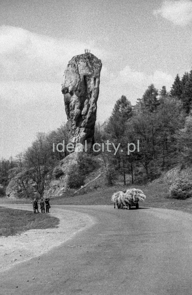 Pionowe ujęcie na pionową skałę o kształcie maczugi, której przygląda się trzech mężczyzn w płaszczach. Wzdłuż drogi jedzie wóz z sianem