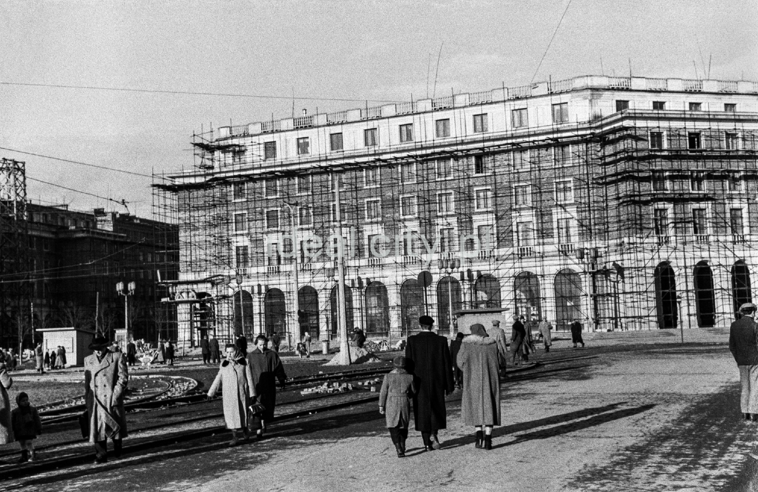 Ludzie spacerują po niewykończonym placu miejskim, otoczonym monumentalną zabudową mieszkalną.